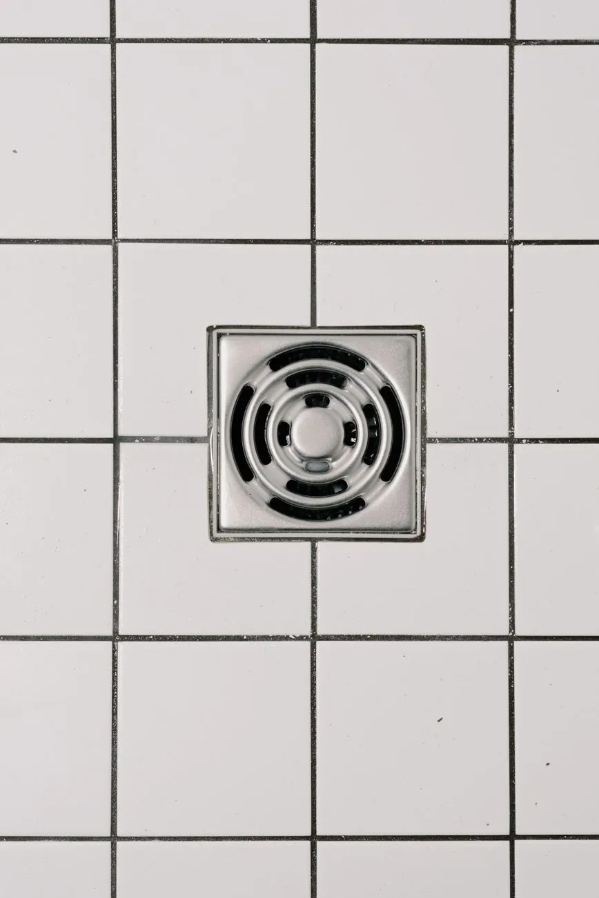 A square stainless steel floor drain set into white tiles